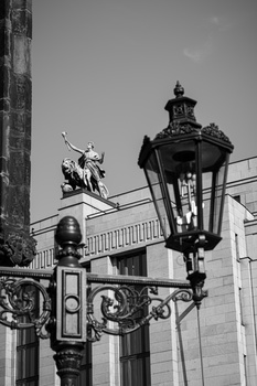 Ornate street lamp with decorative metalwork and a statue visible in the background against a classical building.