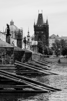 Black and white photograph of a Gothic tower and bridge with wooden logs floating in water below.