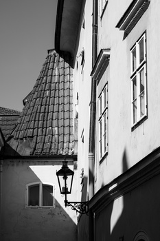 Black and white architectural photograph of a narrow European building corner with tiled roof and wall-mounted lantern.