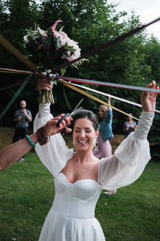 Bride in white off-shoulder dress holding bouquet with arms raised, smiling at outdoor wedding reception.