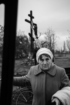 Elderly woman in winter coat and headscarf gazes at ornate cross in bare landscape.