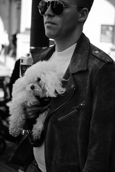 Man in dark leather jacket and sunglasses holding a small white dog outdoors.