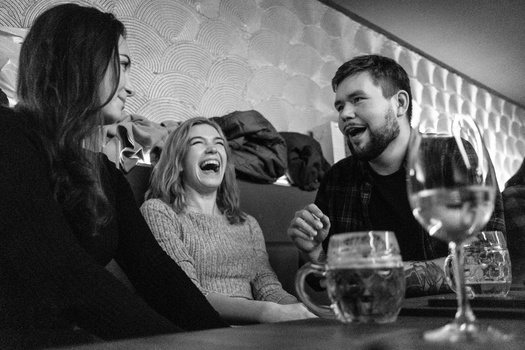 Three people laughing together at a table with glasses of water and wine, shot in black and white.