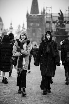 Two women walking toward camera on a bridge with Gothic architecture and other pedestrians in soft-focus background.