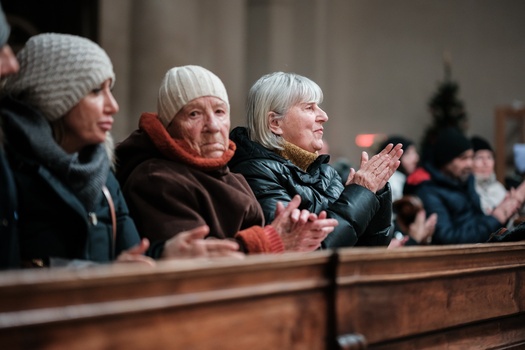 Three older women seated in a pew, wearing winter coats and hats, clapping with focused expressions.