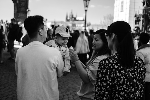 Family with young child in light-colored cap standing among crowds on a cobblestone street in a European city center.