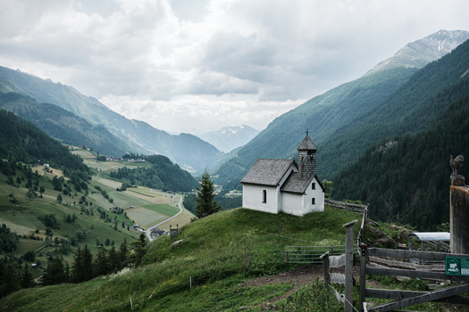 White chapel with steeple perched on a grassy hillside overlooking an Alpine valley.