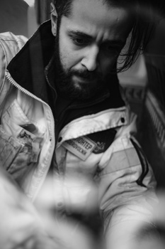 Bearded man in denim shirt and dark jacket looking down at his hands in black and white portrait.