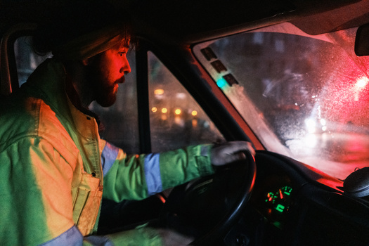 Driver inside vehicle at night illuminated by red and blue emergency lights reflecting through windshield.
