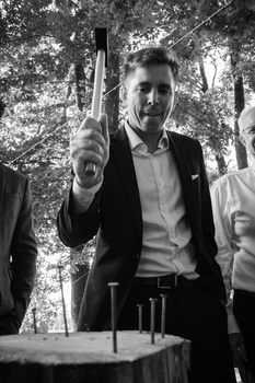 Man in blazer and white shirt holding an axe overhead among trees in black and white photography.