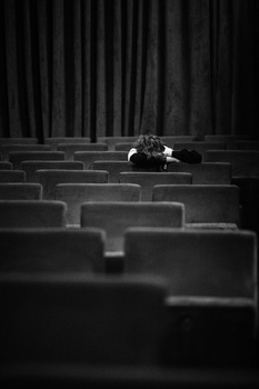 A lone figure sits in an empty theater surrounded by rows of vacant seats.