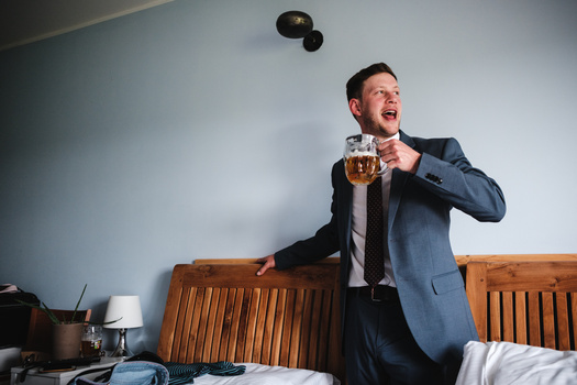 Man in business suit standing on bed, holding beer mug and looking upward with expression of joy.