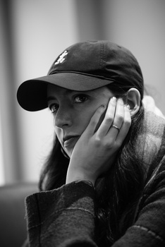 Black and white portrait of a person wearing a baseball cap and dark jacket, hand raised to face in a contemplative pose.