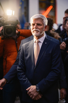 Older man in navy suit and glasses at a public event with camera operators and crowds in background.