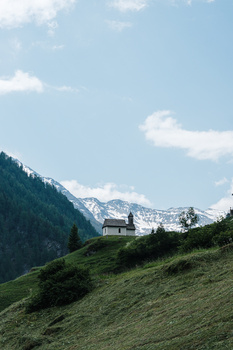 Alpine chalet nestled on a green hillside with snow-capped mountains in the background under a partly cloudy sky.