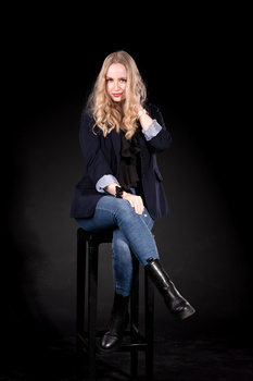 Woman in dark blazer and jeans sitting on black stool against black background.