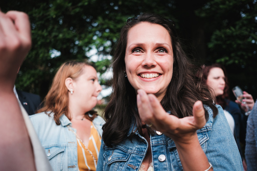 Woman in denim shirt smiling at camera during outdoor daytime gathering with blurred people behind her.