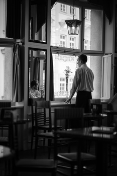 Man standing at large window in monochrome interior, looking out at urban buildings.
