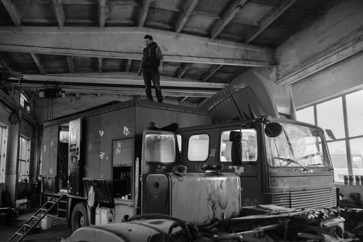 Black and white photograph of an abandoned vintage trailer in an industrial garage with a man standing on its roof.