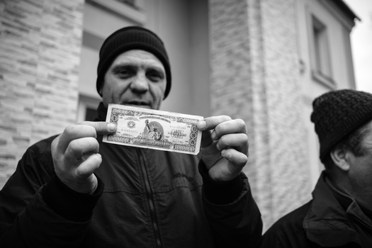 Man in dark winter clothing holding a dollar bill toward the camera against a brick building backdrop.