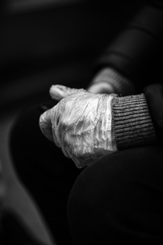 Elderly hands wearing knitted gloves held together against dark clothing in black and white photography.