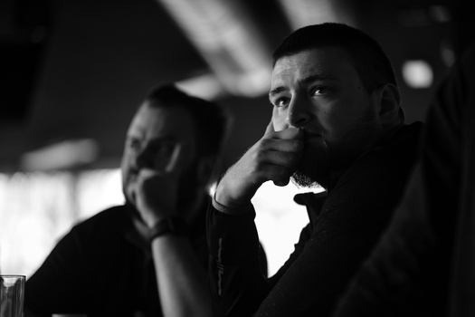 Two men in contemplative poses shot in black and white, with the man in foreground resting his chin on his hand.