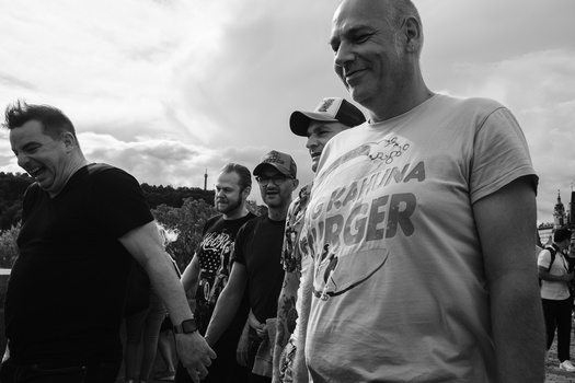 Group of people gathered outdoors in black and white photography, with a man in a graphic t-shirt smiling in the foreground.