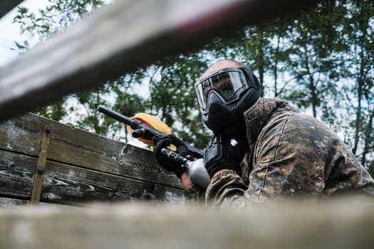 Paintball player in protective gear aiming marker gun behind wooden barrier.