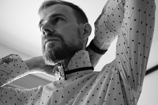 Bearded man in polka-dot shirt looking upward with arms raised in contemplative pose.