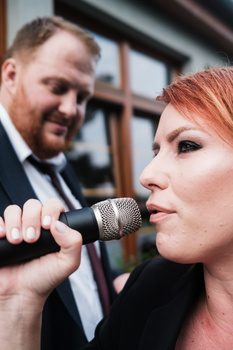 Woman with reddish hair singing into a microphone at an event, with a man in a suit visible in the background.