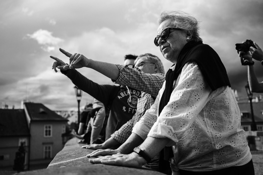 Elderly woman in sunglasses pointing toward horizon while sitting with younger person against cloudy sky.