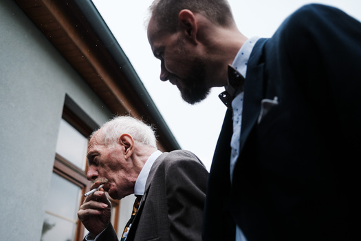 Two men in conversation, one elderly in dark suit and one younger in navy blazer, photographed outdoors against a light building facade.