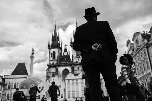 Man in dark suit and fedora hat standing in a European town square with Gothic church spires in the background.