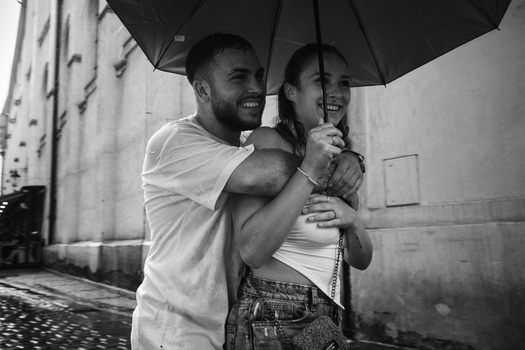 Man and woman smiling while holding an umbrella together in a rainy urban setting.