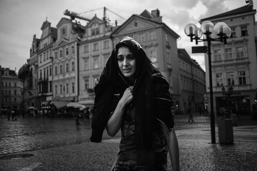 Woman in dark hooded cloak in a European town square during overcast conditions.