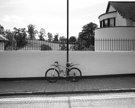 Bicycle leaning against a pole in front of a white wall with modernist architecture visible behind a fence.