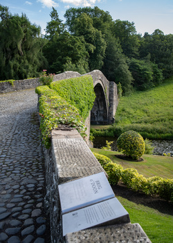 Stone archway covered in moss overlooking a manicured garden with green lawn and trees.