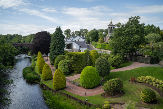 Riverside estate with manicured gardens, brick pathways, and church spire visible through mature trees.