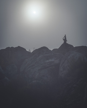 Silhouetted figure standing atop a rocky mountain peak against a bright, hazy sky.