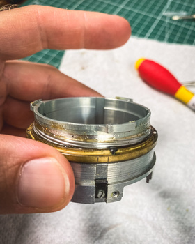 Close-up of a disassembled lens element or optical component held in hands, showing metal and glass parts.