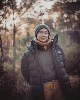 Person wearing a gray beanie and black puffer jacket standing in a wooded area during golden hour.