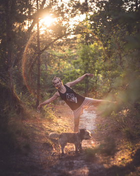 Dancer performing a high kick on a forest path with golden sunlight filtering through trees.