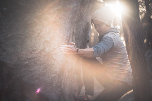 Person in casual clothing reaching toward a textured concrete wall, backlit by bright sunlight creating lens flare and atmospheric glow.
