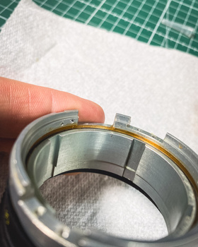 Close-up of a person's finger holding a metal ring with a gold band detail against a white fabric and green textured background.