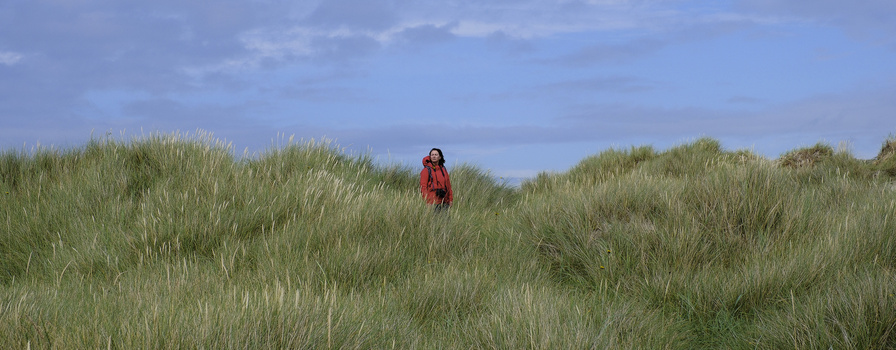 Woman in red jacket standing among tall beach grasses under blue sky.