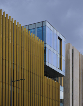 Modern building facade with mustard-yellow vertical panels and blue-glass protruding window modules against overcast sky.