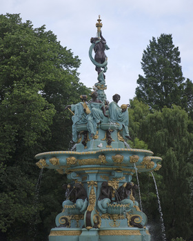 Ornate turquoise and gold fountain with sculptural figures, surrounded by green trees under cloudy sky.