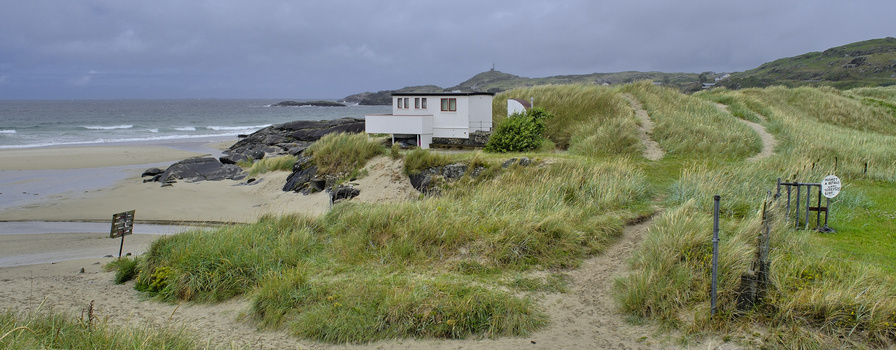 White modern house perched on grassy sand dunes overlooking a rocky beach and ocean.