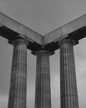 Three fluted classical columns supporting a concrete beam structure, photographed from below against an overcast sky.