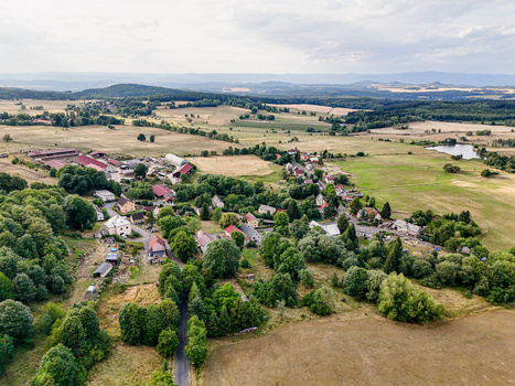 Aerial view of a rural village nestled among green fields and forests beneath an overcast sky.